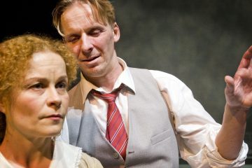 Etualalla on nainen, jolla on kiharat hiukset ja joka näyttää vakavalta, kun taas hänen takanaan on mies, jolla on valkoinen paita, liivi ja punainen solmio ja joka näyttää intensiiviseltä dramaattisessa kohtauksessa.