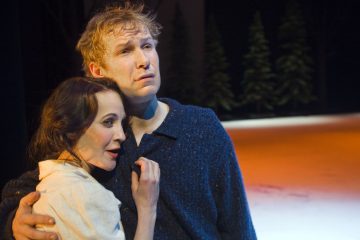 Nainen ja mies seisovat yhdessä syleillen lavalla. Nainen näyttää huolestuneelta ja pitelee miehen rintaa, kun taas mies katsoo mietteliäästi eteenpäin. Taustalla näkyy lunta ja mäntyjä.