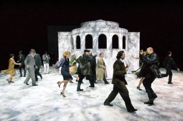 Joukko tyylikkäisiin vintage-vaatteisiin pukeutuneita ihmisiä liikkuu tarmokkaasti lavalla suuren valkoisen, pyöreän, kaarevilla aukoilla varustetun rakennuksen edessä dramaattisessa valaistuksessa, joka heittää varjoja lattiaan ja seiniin.