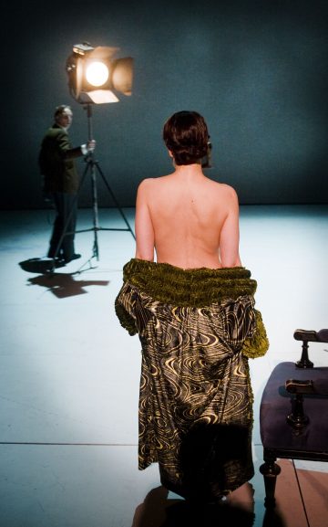 Lyhyet tummat hiukset omaava henkilö seisoo lavavalojen alla takaapäin ja paljain selin, ja hänen vyötärönsä ympärille on puettu kuvioitu vaate. Taustalla toinen henkilö seisoo suuren valonheittimen vieressä.