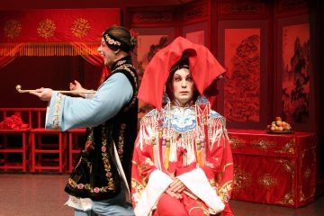 Kaksi perinteiseen kiinalaiseen oopperapukuun pukeutunutta esiintyjää, joista toinen istuu etualalla punaisessa puvussa ja päähineessä ja toinen siniseen ja mustaan pukeutuneena seisoo takana, ja taustalla on koristeltu punainen tausta, jossa on kiinalaisia huonekaluja ja taittuvia näyttöjä.