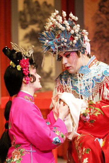 Kaksi esiintyjää koristeellisissa perinteisissä kiinalaisissa oopperapuvuissa ja -meikeissä pitävät toisiaan kädestä näyttämöllä ja ilmaisevat voimakkaita tunteita. Kohtaus on värikkäiden vaatteiden, taidokkaiden päähineiden ja maalatun taustan värittämä.