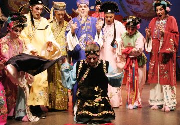 Ryhmä näyttelijöitä värikkäissä, koristeellisissa perinteisissä kiinalaisissa oopperapuvuissa esiintyy lavalla; yksi näyttelijä polvistuu dramaattisesti keskellä, kun muut katsovat sitä ilmeikkäin kasvoin.