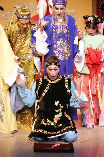 Värikkääseen, perinteiseen kiinalaiseen oopperapukuun pukeutunut esiintyjä polvistuu lavalla yllättynyt ilme kasvoillaan, ja hänen ympärillään on kolme muuta hienosti pukeutunutta näyttelijää, jotka kaikki reagoivat dramaattisesti. Koristeellinen laatikko makaa avoimena lattialla.