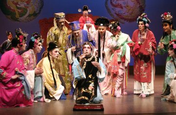 Ryhmä värikkäisiin perinteisiin kiinalaisiin oopperapukuihin pukeutuneita esiintyjiä kerääntyy lavalle mustaan ja kultaan pukeutuneen keskushahmon ympärille, joka poseeraa dramaattisesti näyttämön valaistuksen alla. Koristeelliset päähineet ja kirjaillut kaavut ovat näkyvissä.