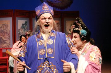 Lavalla esiintyy näyttelijöitä perinteisissä kiinalaisissa oopperapuvuissa, joista yksi siniseen kaapuun ja hattuun pukeutunut näyttelijä näyttää yllättyneeltä tai vihaiselta ja toinen vaaleanpunaiseen pukuun pukeutunut näyttelijä näyttää huutavan hänelle. Taustalla on koristeellisia valkokankaita.