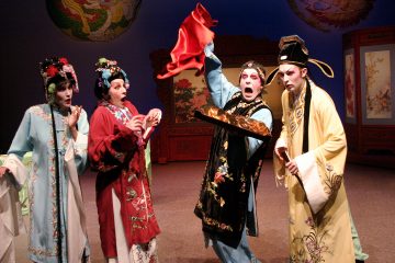 Lavalla esiintyy neljä näyttelijää värikkäissä, koristeellisissa perinteisen kiinalaisen oopperan puvuissa. Yksi näyttelijä nostaa dramaattisesti punaista liinaa, ja muut reagoivat siihen yllättyneinä ja ilmeikkäinä. Koristeelliset näytöt ja maalatut kulissit koristavat näyttämöä.