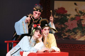 Kolme perinteiseen kiinalaiseen oopperaan pukeutunutta ja meikattua esiintyjää poseeraa dramaattisesti. Kaksi nojaa punaiseen pöytään, ja kolmas seisoo heidän takanaan ja tekee eleitä. Taustalla on kukkamaalaus.