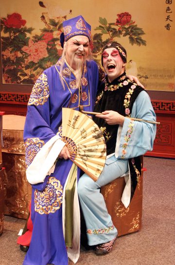 Kaksi perinteiseen kiinalaiseen oopperapukuun ja meikkiin pukeutunutta esiintyjää hymyilee ja poseeraa yhdessä sisätiloissa. Toisella on sininen kaapu ja hattu, ja hänellä on kädessään viuhka, kun taas toisella on vaaleansininen ja musta kaapu, ja hän istuu kultaisen arkun päällä. Heidän takanaan on kukkatausta.