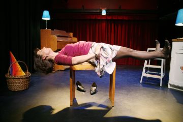 Vaaleanpunaiseen yläosaan ja kukkahameeseen pukeutunut nainen makaa vaakasuorassa selällään kahden tuolin välissä tasapainoillen, jalat ja hartiat tuettuina, kengät lattialla hänen alapuolellaan. Näyttämö on lämpimästi valaistu rekvisiitan ja taustalla olevien punaisten verhojen avulla.