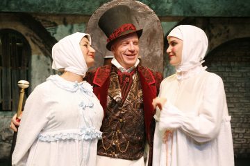 Kolme teatteripukuista näyttelijää seisoo yhdessä; silinterihattuinen ja koristeelliseen punaiseen takkiin pukeutunut mies hymyilee kahden valkoisiin päähineisiin ja pukuihin pukeutuneen naisen välissä, jotka katsovat häntä. Tausta muistuttaa vanhaa, maalaismaista katukuvaa.