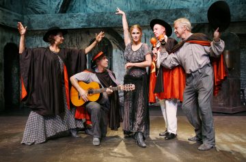 Viisi näyttelijää lavalla puvuissa esittää dramaattisen kohtauksen. Yksi soittaa kitaraa istuen, kun muut poseeraavat vilkkaasti, yksi nainen nostaa kättään ja muut elehtivät ilmeikkäästi. Tausta viittaa vanhaan rakennukseen.