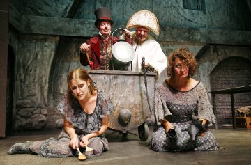 Kaksi harmaisiin mekkoihin pukeutunutta naista istuu lattialla surullisen näköisinä, kengät kädessä. Heidän takanaan hymyilee kaksi teatteriasuihin pukeutunutta miestä, joista toisella on punainen hattu ja toisella valkoinen ja suuri naamio, ja he seisovat näyttämöllä suuren metalliastian vieressä.