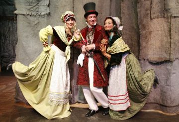 Kolme viktoriaanisen ajan pukuihin pukeutunutta aikuista poseeraa yhdessä lavalla. Keskellä olevalla miehellä on punainen kuvioitu takki ja korkea musta hattu, ja kahdella naisella molemmin puolin on päähineet ja pitkät mekot, joissa on esiliina.