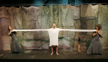 Lavalla seisoo kolme näyttelijää. Keskellä olevalla miehellä on pitkä valkoinen paita, jonka hihat on ojennettu ja jota kaksi kuviomekkoon pukeutunutta naista pitelee molemmin puolin. Taustalla on kuvioitu, verhottu kangas.