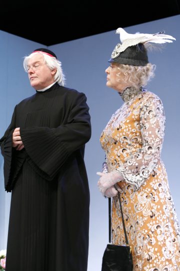 Mustaan kaapuun pukeutunut vanhempi mies ja kukkamekkoon pukeutunut nainen, jolla on pitsikäsineet ja valkoisen kyyhkyn koristama hattu, seisovat lavalla vierekkäin ja näyttävät vakavilta.