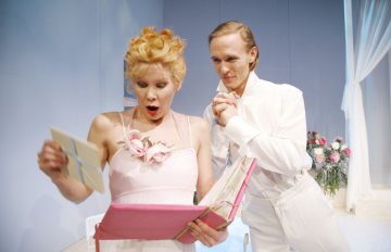 Vaaleanpunaiseen mekkoon pukeutunut nainen näyttää yllättyneeltä pitäessään kädessään suurta vaaleanpunaista kirjaa ja kirjettä, kun valkoiseen pukuun pukeutunut mies seisoo hänen vieressään hymyillen ja kätensä yhteen kietoen kirkkaasti valaistussa huoneessa, jonka taustalla on kukkia.