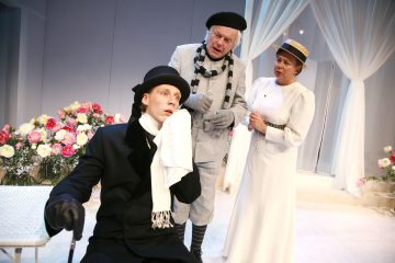 Lavalla esiintyy kolme aikakauden pukuihin pukeutunutta näyttelijää: yksi istuu penkillä liina suullaan ja näyttää dramaattiselta, kun taas kaksi muuta seisovat lähellä ja vaikuttavat huolestuneilta. Taustaa koristavat kukat ja valkoiset verhot.