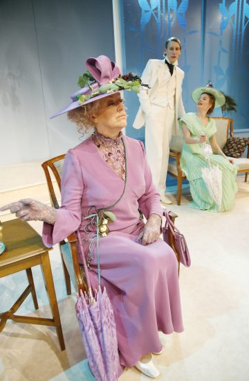 Kolme näyttelijää lavalla aikakauden puvuissa: istuva vanhempi nainen laventelinvärisessä mekossa ja hienostuneessa hatussa, nainen mintunvihreässä mekossa ja aurinkovarjo kädessään sekä seisova henkilö valkoisessa puvussa. Taustalla sinipaneloitu tausta.