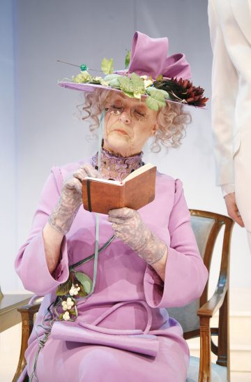 Iäkäs nainen, jolla on laventelinvärinen mekko ja siihen sopiva hattu, jossa on kukkia ja lehtiä, istuu lukemassa pientä kirjaa puisella tuolilla valoisassa huoneessa pyöreissä silmälaseissa ja pitsikäsineissä.