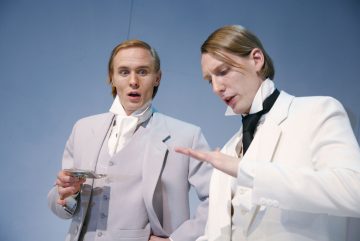 Kaksi valkoisiin ja vaaleanharmaisiin pukuihin pukeutunutta miestä seisoo sinistä taustaa vasten. Toinen näyttää yllättyneeltä pitelemässä taskukelloa, ja toinen elehtii kädellään ja näyttää selittävän jotain.