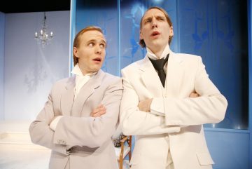 Kaksi vaaleisiin juhlapukuihin pukeutunutta ihmistä seisoo vierekkäin kädet ristissä eri suuntiin katsoen kirkkaasti valaistussa huoneessa, jonka seinät ovat siniset ja jonka taustalla on kattokruunu.