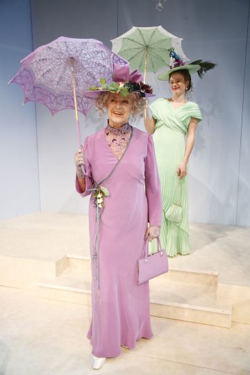Kaksi vintage-tyyliseen mekkoon ja hattuun pukeutunutta naista seisoo vaalealla näyttämöllä, kummallakin koristeellinen aurinkovarjo kädessään. Edessä olevalla naisella on laventelinvärinen asu, takana olevalla naisella vaaleanvihreä. Molemmat hymyilevät.