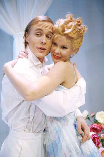 Tyylikkääseen, vintage-tyyliseen valkoiseen vaatetukseen pukeutuneet mies ja nainen syleilevät ja hymyilevät. Taustalla on valkoisia verhoja ja kukkia, jotka luovat romanttisen, teatraalisen tunnelman.