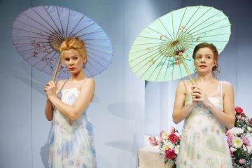 Kaksi kevyeen kukkamekkoon pukeutunutta naista pitää koristeellisia aurinkovarjoja olkapäidensä takana. He seisovat kukkakimpun vieressä vaaleansinistä taustaa vasten ja näyttävät esiintyvän lavalla.