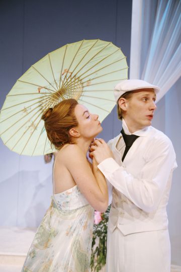 Kukkamekkoon pukeutunut nainen pitelee vihreää aurinkovarjoa ja nojaa valkoiseen pukuun ja lippikseen pukeutunutta miestä kohti; molemmat näyttävät näyttelevän näyttämöllä, jossa on pehmeä sinivalkoinen tausta.