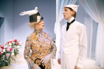 Vanhempi nainen, jolla on kuvioitu mekko ja hattu, jonka päällä on valkoinen lintu, puhuu valkoiseen pukuun ja lippikseen pukeutuneelle nuorelle miehelle. He seisovat sisätiloissa kukkien ja valkoisten verhojen ympäröimänä.