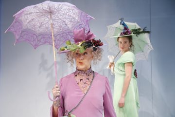 Kaksi hienostuneisiin pukuihin pukeutunutta naista seisoo vaaleansinistä taustaa vasten. Edessä olevalla naisella on hieno vaaleanpunainen mekko ja hattu, ja hänellä on kädessään vaaleanpunainen pitsinen aurinkovarjo. Takana olevalla naisella on vaaleanvihreä päällinen, ja hänellä on valkoinen aurinkovarjo ja kuppi.