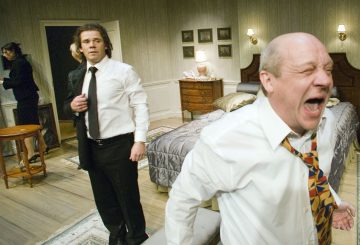 Etualalla oleva mies huutaa suu auki, värikäs solmio vinossa. Hänen takanaan seisoo toinen valkoiseen paitaan ja mustaan solmioon pukeutunut mies huolestuneen näköisenä makuuhuoneessa, jonka taustalla on kaksi muuta ihmistä.