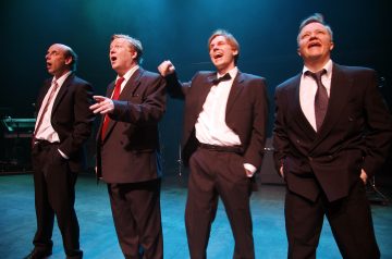 Neljä mustiin pukuihin ja solmioihin pukeutunutta miestä seisoo näyttämöllä dramaattisessa valaistuksessa, ja he näyttävät laulavan tai esiintyvän innokkaasti, ja jotkut heistä elehtivät tai hymyilevät. Taustalla on musiikkilaitteita ja sini-violetti hehku.