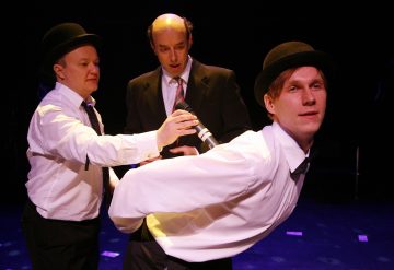 Lavalla esiintyy kolme pukuihin ja keilahattuihin pukeutunutta miestä. Yksi mies kumartuu eteenpäin, kun toinen pitää mikrofonia selällään, ja kolmas mies katselee takaapäin dramaattisessa valaistuksessa.