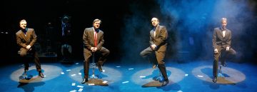 Neljä pukumiestä istuu jakkaroilla valonheittimien alla pimeällä, savuisella näyttämöllä. Lattialle on ripoteltu paperinpaloja. Tausta on hämärä, ja hahmoja ja esineitä näkyy hädin tuskin.