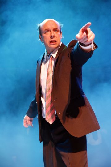 Pukuun ja solmioon pukeutunut mies seisoo dramaattisessa sinisessä valaistuksessa ja osoittaa eteenpäin vakava ilme kasvoillaan. Hänen ympärillään on sumua tai savua, joka luo teatraalisen tunnelman.