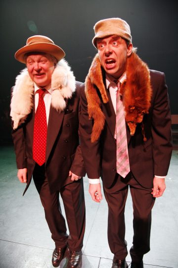 Lavalla seisoo vierekkäin kaksi pukuihin ja hattuihin pukeutunutta miestä, joilla kummallakin on kaulassaan turkistakki. Toisella miehellä on punainen solmio ja hän nauraa silmät kiinni, kun taas toisella on vaaleanpunainen solmio ja hän näyttää suurisilmäiseltä ja eloisalta.