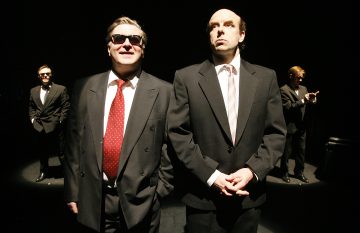 Viisi pukumiestä seisoo pimeällä näyttämöllä. Kaksi on edessä - yhdellä on aurinkolasit ja punainen solmio, toinen katsoo ylöspäin. Taustalla on kolme miestä, kaksi keskustelemassa ja yksi seisoo yksin.