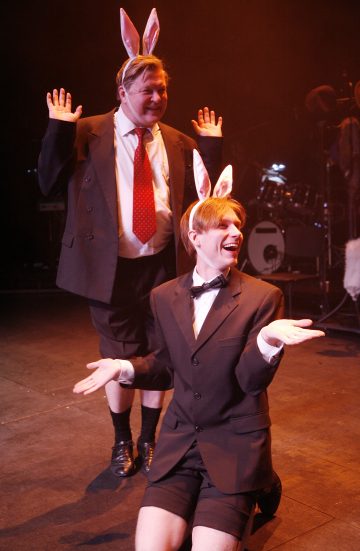 Lavalla esiintyy kaksi pukuihin ja pupunkorviin pukeutunutta ihmistä; toinen seisoo kädet ylhäällä, toinen polvistuu kädet ojennettuina, ja molemmat hymyilevät innostuneesti. Taustalla näkyy soittimia.