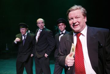 Lavalla seisoo neljä pukumiestä dramaattisessa valaistuksessa. Edessä oleva mies hymyilee ja pitää kädessään rahaa, kun taas kolme takana olevaa miestä, joista kahdella on hattu, näyttävät vakavilta, mikä viittaa teatteriesitykseen tai kohtaukseen.