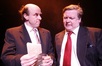 Kaksi pukumiestä seisoo lähekkäin. Toisella on kädessään juoma ja taiteltu sanomalehti, ja hän katsoo toista sivusuunnassa. Toinen mies, jolla on punainen solmio, katsoo lehteä alaspäin. Tausta on tumma ja pehmeästi valaistu.