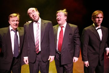 Lavalla seisoo neljä tummiin pukuihin pukeutunutta miestä dramaattisessa valaistuksessa. Heillä on ilmeikkäät kasvonilmeet ja kehonkieli, jotka viittaavat siihen, että he esittävät tai näyttelevät komedia- tai teatterikohtausta.