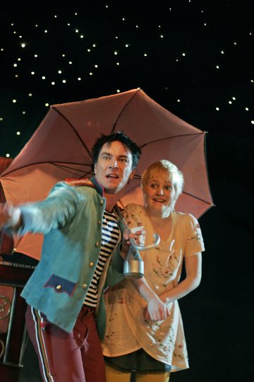 Lavalla kaksi ihmistä seisoo lähekkäin punaisen sateenvarjon alla, toinen osoittaa dramaattisesti eteenpäin. Taustalla on pimeää ja pieniä, tähdenmuotoisia valoja. Molemmilla on ilmeikkäät kasvot ja teatteripuvut.