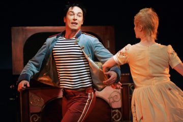 Lavalla esiintyy kaksi näyttelijää, joista toisella on raidallinen paita, sininen takki ja punaiset housut ja toisella vaalea mekko. He ovat kesken tanssin puisen lavastuksen edessä, ja heidän kasvonsa ovat ilmeikkäät ja dynaamiset.