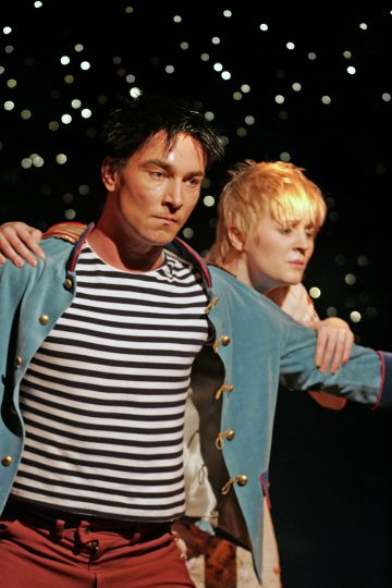 Lavalla kaksi esiintyjää, toinen tummahiuksinen, sininen takki ja raidallinen paita, toinen lyhyet vaaleat hiukset, molemmilla vakavat ilmeet ja kädet ojennettuina. Taustalla välkkyvät valot.