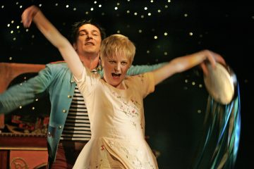 Vaaleaan mekkoon pukeutunut nainen tanssii tarmokkaasti tamburiinin kanssa, ja hänen takanaan seisoo siniseen takkiin ja raidalliseen paitaan pukeutunut mies. Taustalla on tummia, kimaltelevia valoja, jotka luovat eloisan ja teatraalisen tunnelman.