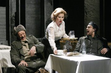 Valkoiseen puseroon pukeutunut nainen tarjoilee juomia kahdelle pöydän ääressä istuvalle sotilaspukuiselle miehelle. Toinen mies nauraa silmät kiinni, kun taas toinen näyttää yllättyneeltä. Ympäristö näyttää olevan hämärästi valaistu kahvila tai ravintola.