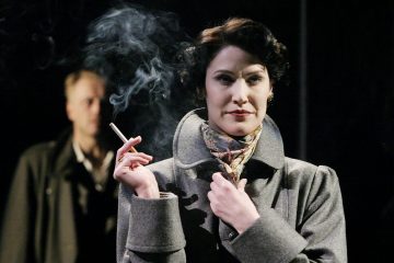 Harmaaseen takkiin ja kukkahuiviin pukeutunut nainen pitää sytytettyä savuketta kädessään ja katsoo luottavaisesti eteenpäin. Taustalla seisoo tummaan takkiin pukeutunut mies, joka on osittain savun peittämä. Asetelma vaikuttaa dramaattiselta ja teatraaliselta.