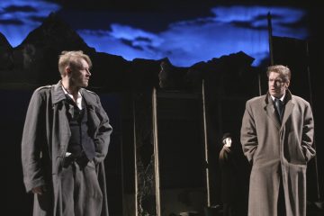 Kaksi pitkiin takkeihin pukeutunutta miestä seisoo vastakkain hämärästi valaistulla näyttämöllä, jonka dramaattinen, rosoinen tausta muistuttaa raunioita; tummiin vaatteisiin pukeutunut henkilö seisoo taustalla sinisen, pilvisen taivaan alla.